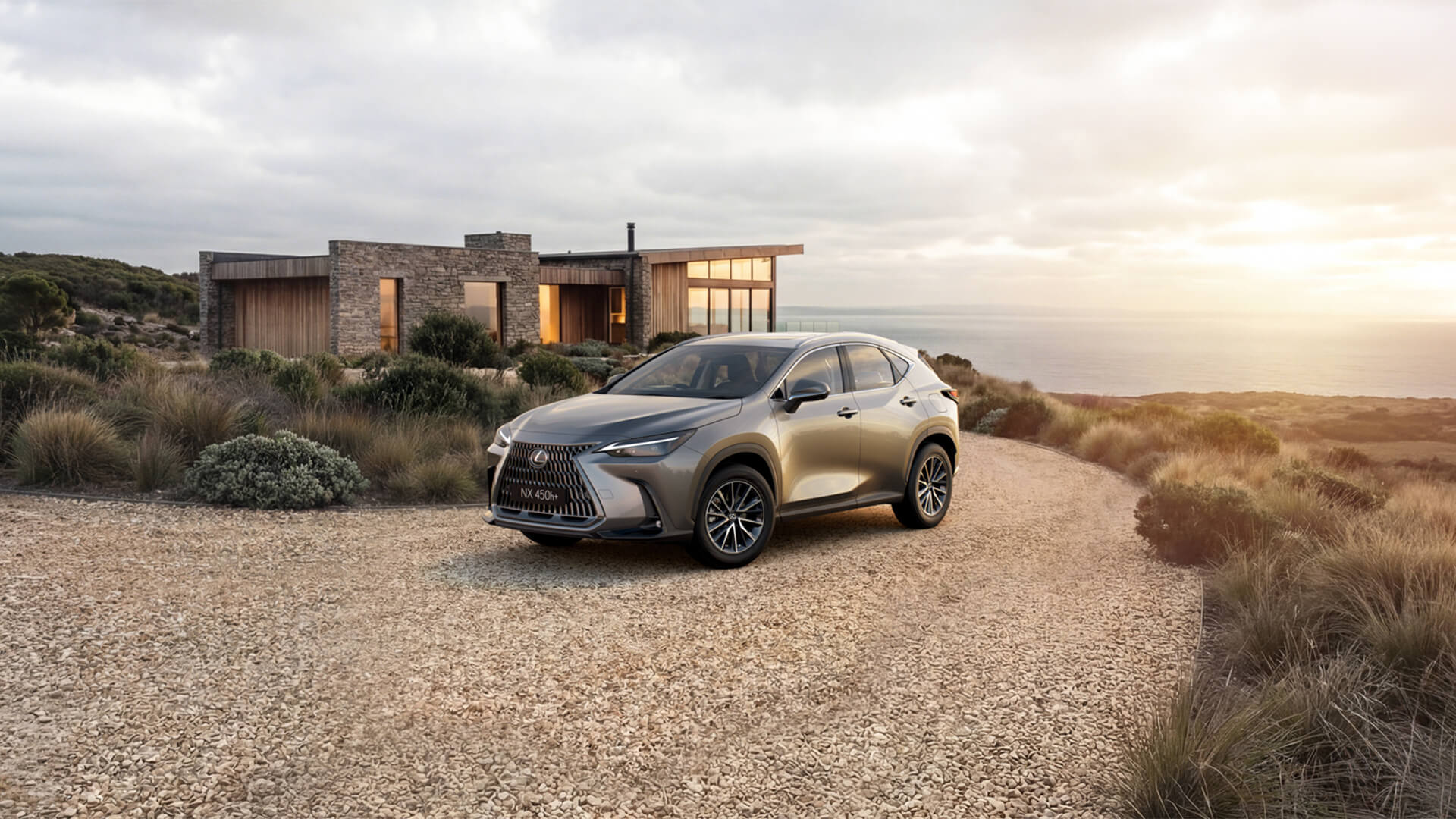 A grey Lexus NX 450h+ SUV parked on a gravel driveway in front of a modern stone and wood house overlooking the ocean during a golden sunset.
