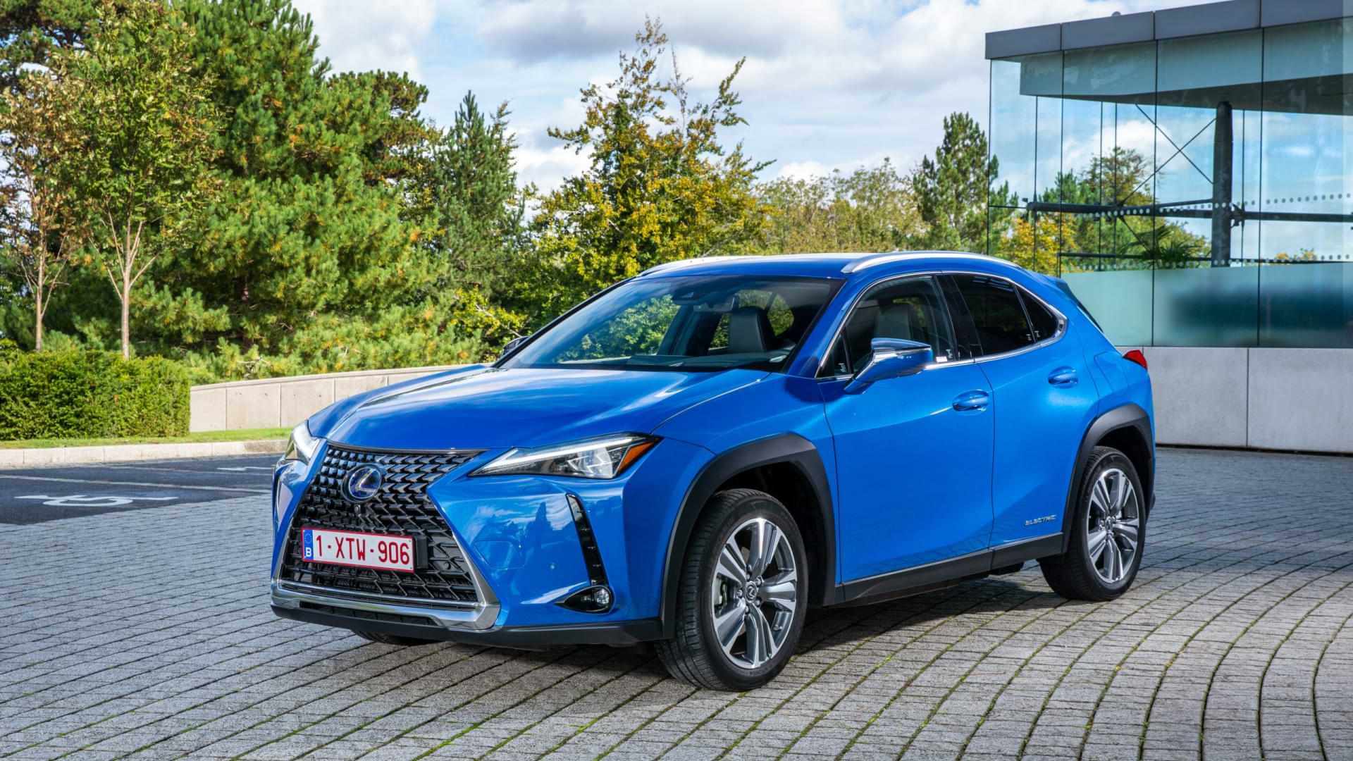An electric blue Lexus UX 300e sits in the driveway of a glass house behind it.